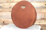 A brown, round leather handbag with handle sits on a white surface against a rustic wooden background.