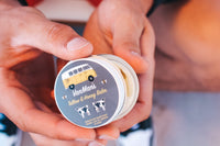 Hands hold open a tin of VanMan's Tallow & Honey Balm, a natural skincare product featuring a VW bus and cows on its label.
