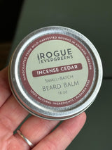 A hand holds a tin of Rogue Evergreens Incense Cedar beard balm made with natural ingredients.
