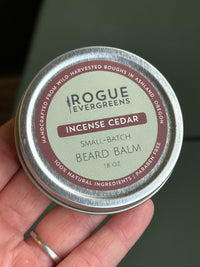 A hand holds a tin of Rogue Evergreens Incense Cedar beard balm made with natural ingredients.
