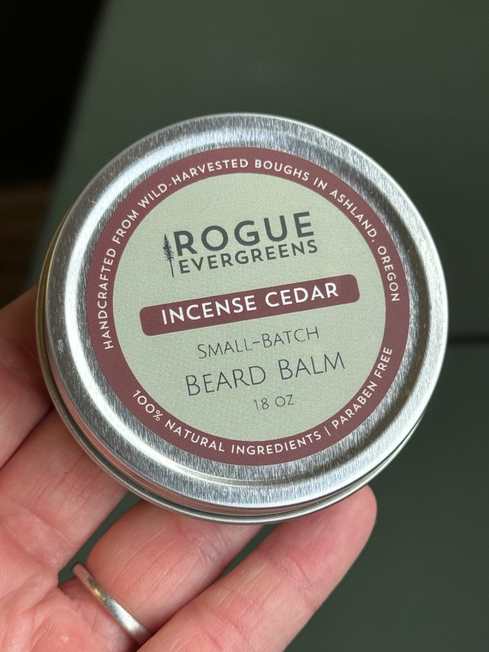 A hand holds a tin of Rogue Evergreens Incense Cedar beard balm made with natural ingredients.
