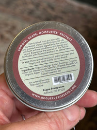 Person holds a Rogue Evergreens beard balm, showcasing its natural ingredient list and promoting smooth, shaped, and moisturized beards.
