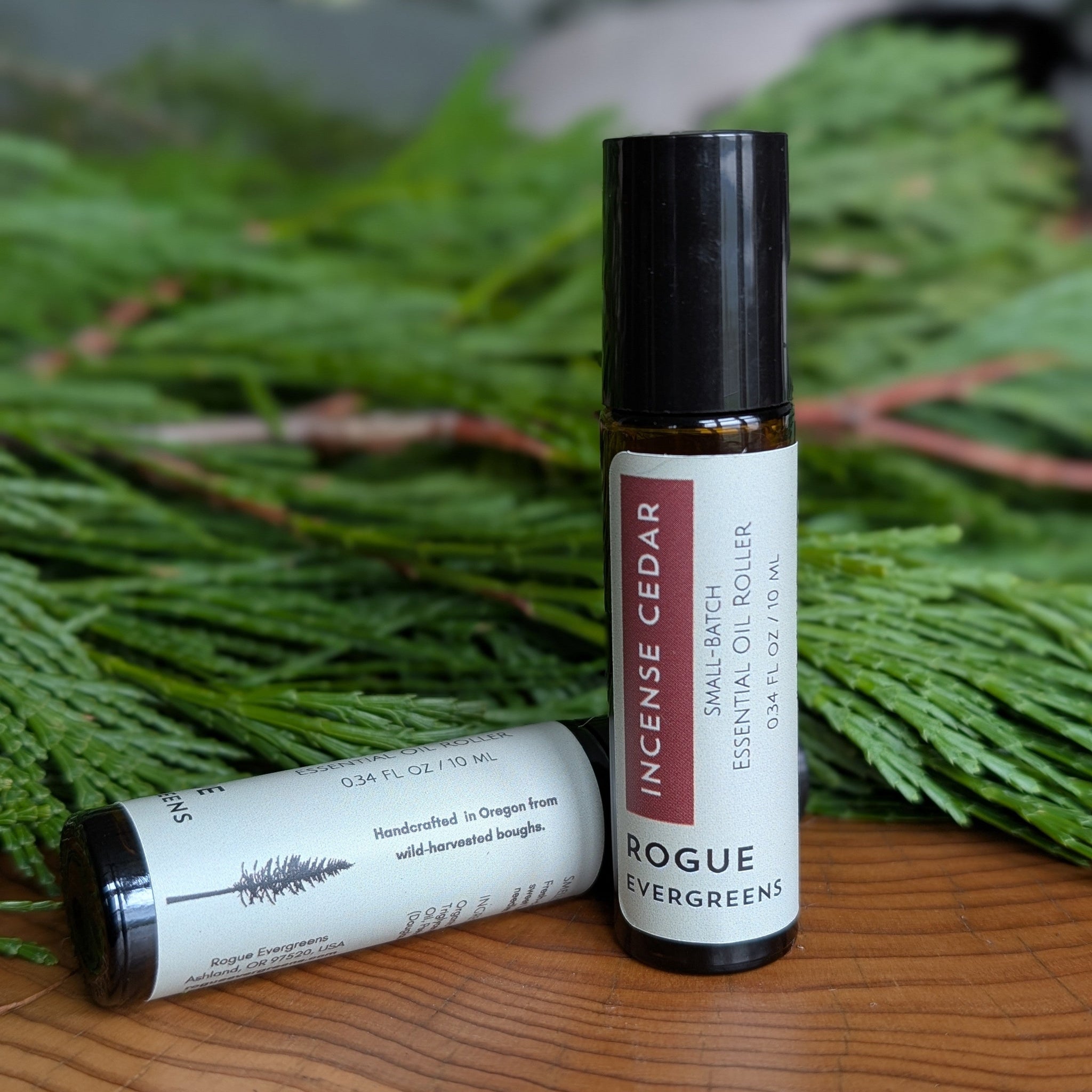 Two Rogue Evergreens incense cedar essential oil roller bottles rest on a wooden surface, accented by evergreen boughs.
