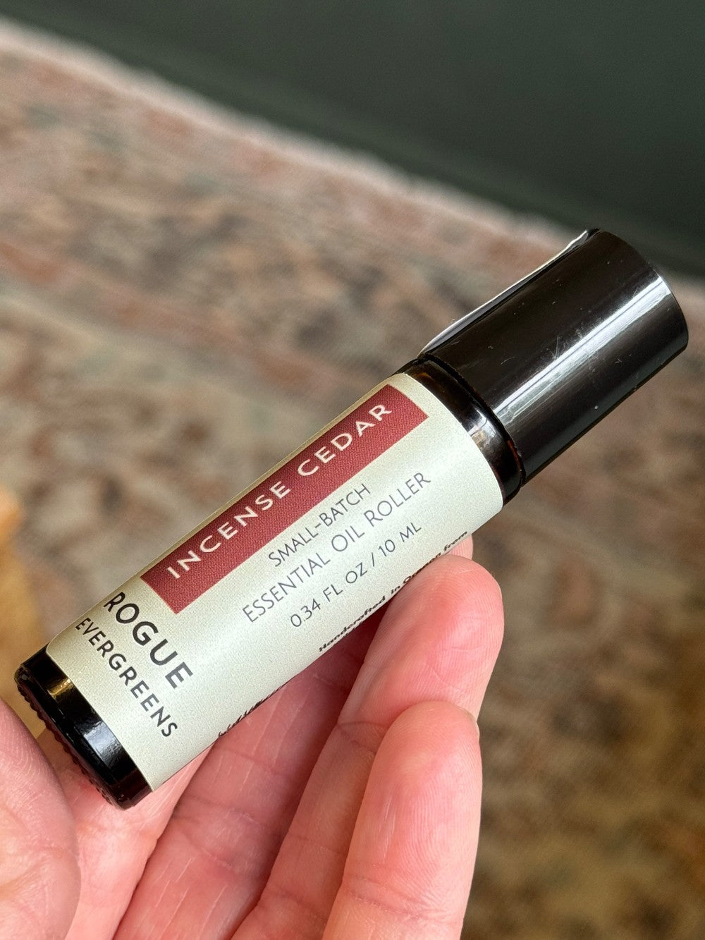 A hand holds a Rogue Evergreens Incense Cedar small-batch essential oil roller bottle.
