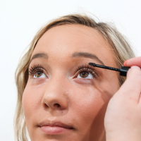 A blonde woman applies black mascara to her upper lashes during a makeup routine.
