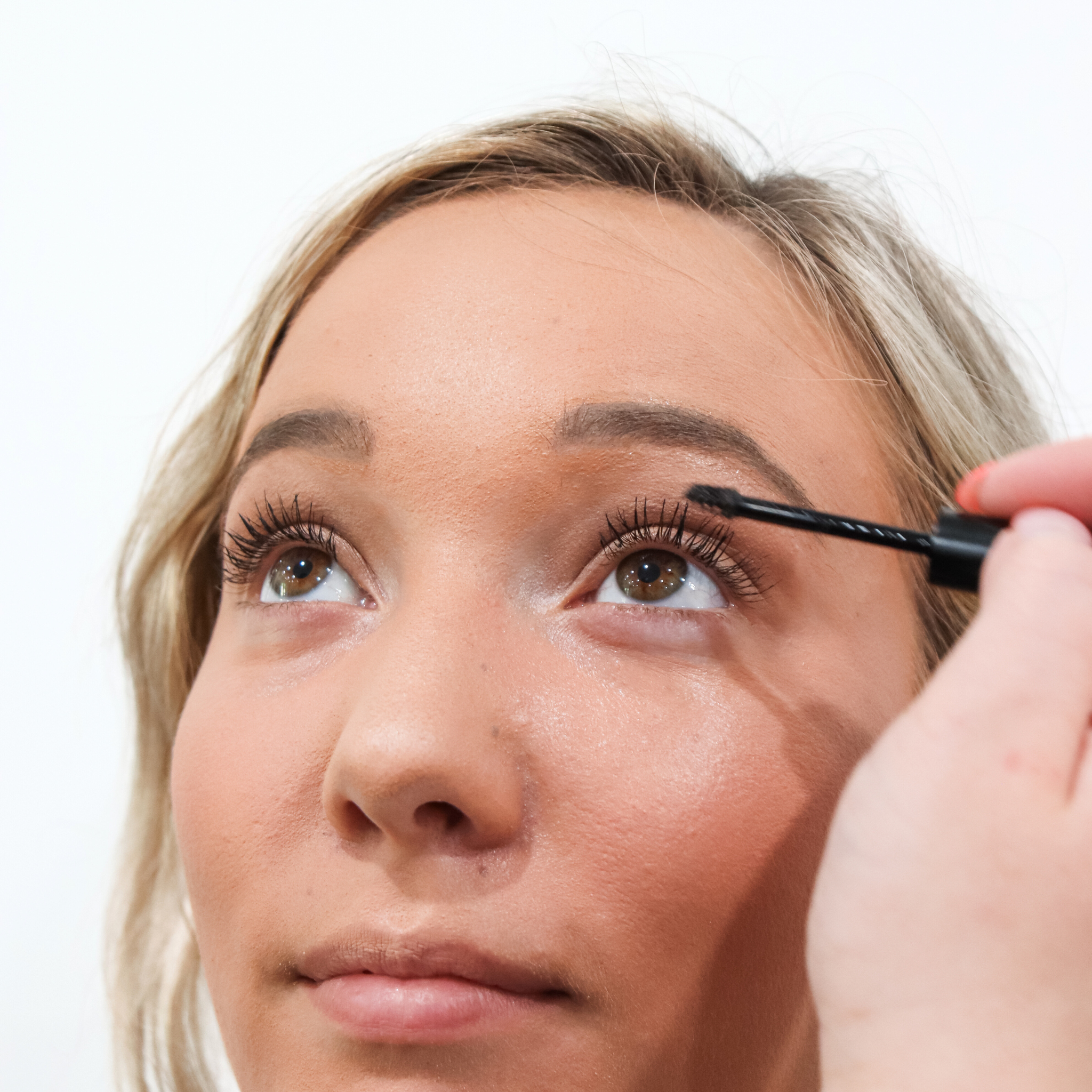 A blonde woman applies black mascara to her upper lashes during a makeup routine.
