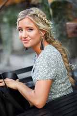 Blonde woman with a braided hairstyle and floral dress looks toward the camera while holding a black tote bag on a bench.