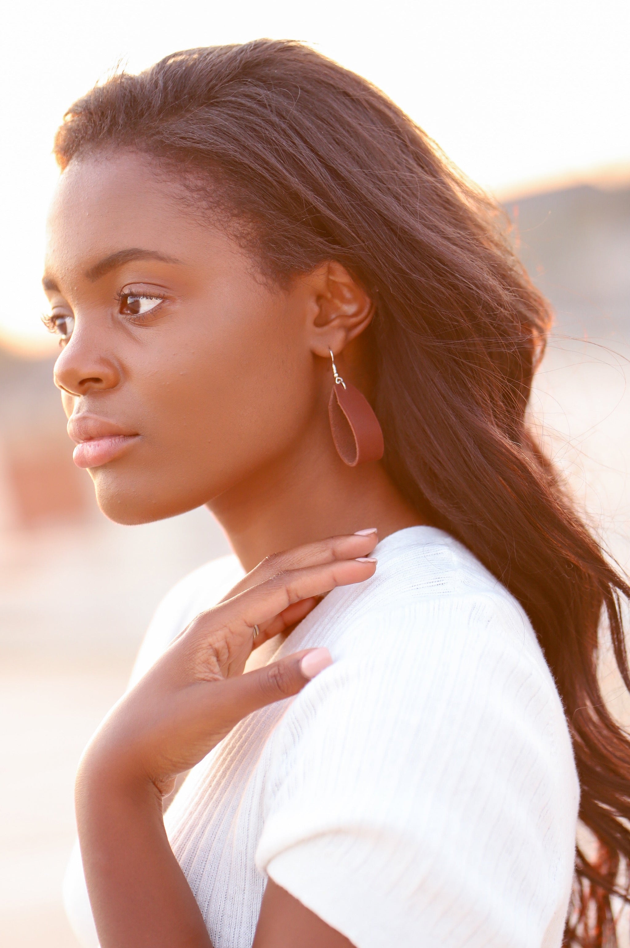 African-American woman models a teardrop-shaped leather earring and a cream-colored sweater, gazing toward a sunlit horizon.