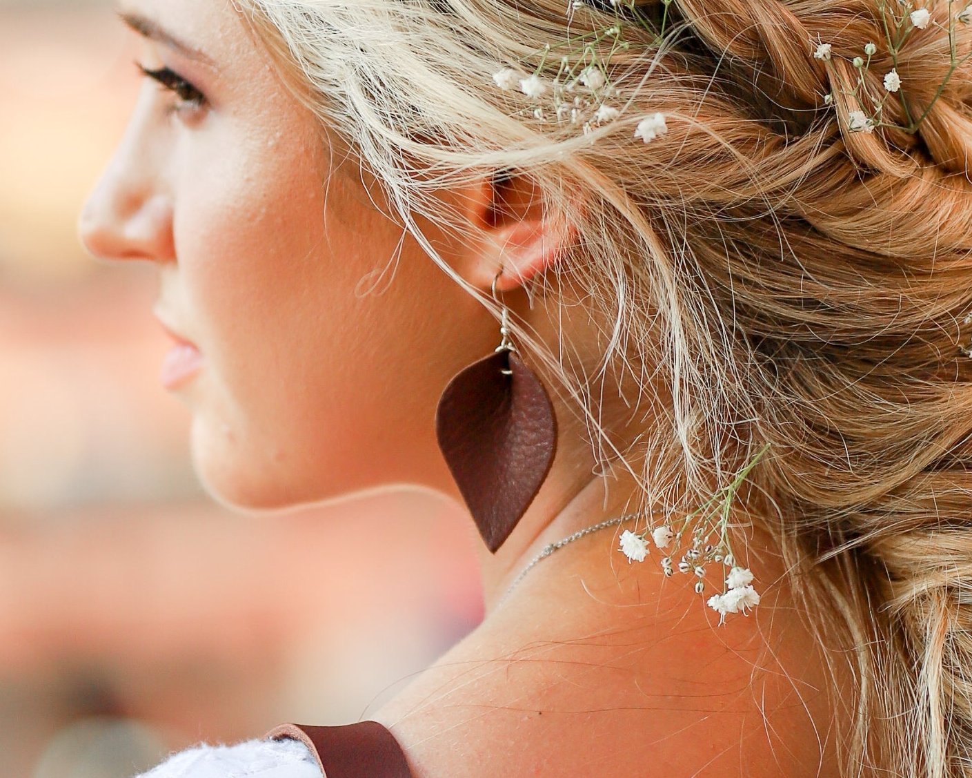 A close-up side profile of a woman with blonde hair adorned with small white flowers, wearing a large dark brown leaf-shaped dangle earring.