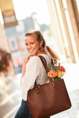 Smiling, young woman carries a brown leather tote bag filled with vibrant, fresh flowers down a sunny street.
