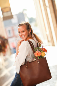 Smiling, young woman carries a brown leather tote bag filled with vibrant, fresh flowers down a sunny street.
