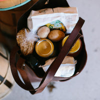 A brown leather tote bag holds groceries including potatoes, granola, canned goods, and a bag of chips.
