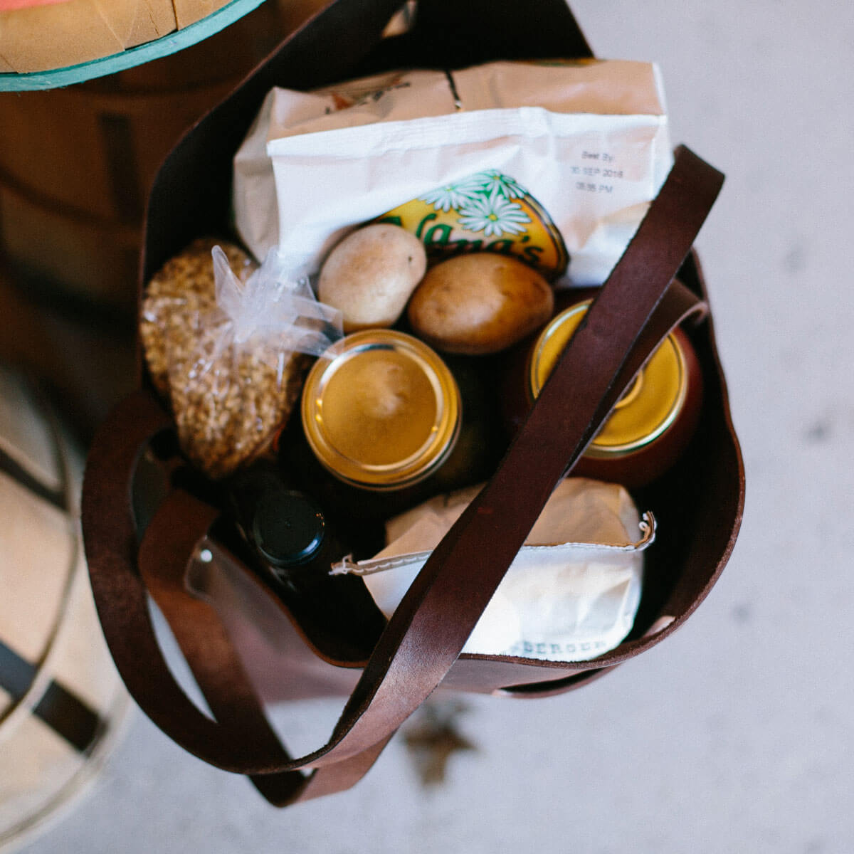 A brown leather tote bag holds groceries including potatoes, granola, canned goods, and a bag of chips.
