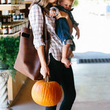A mother carries her baby and a pumpkin while shopping at a farmers market.
