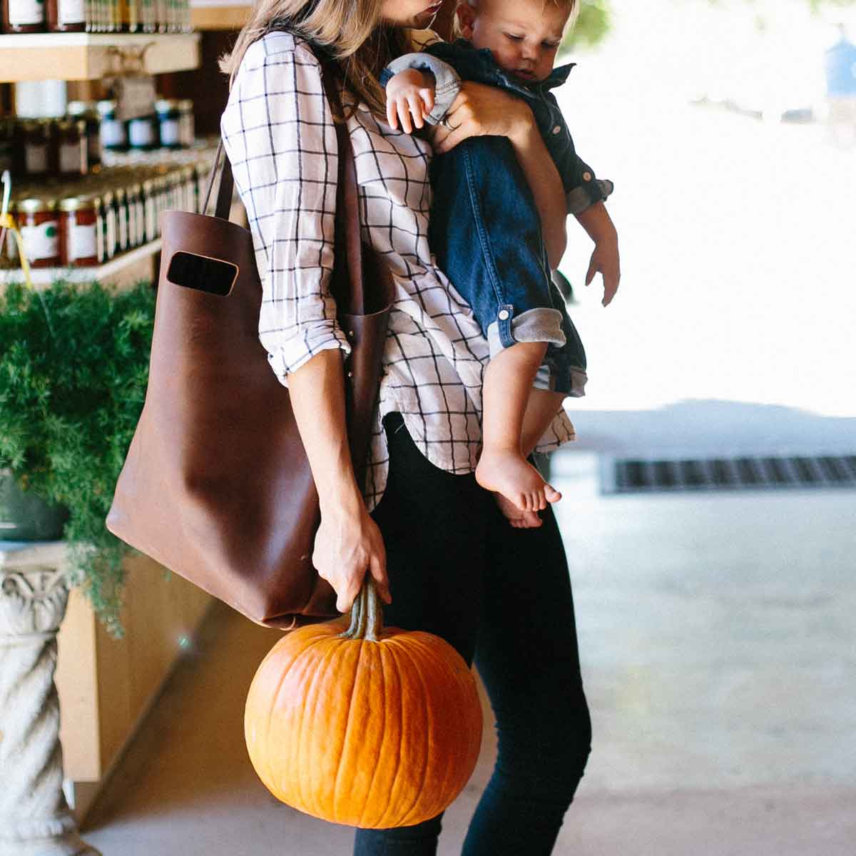 A mother carries her baby and a pumpkin while shopping at a farmers market.
