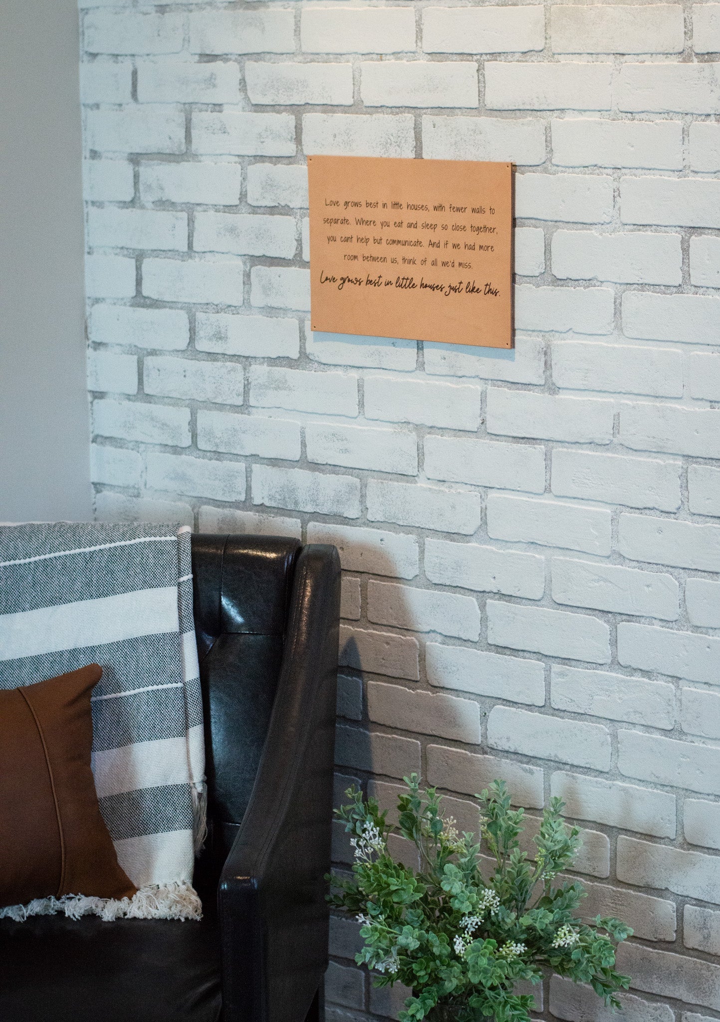 Cozy home decor features a leather chair with a striped blanket and decorative pillow next to a brick wall showcasing a motivational quote art print.
