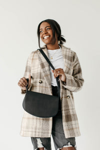 Smiling woman models a black leather crossbody purse and a stylish plaid overcoat.
