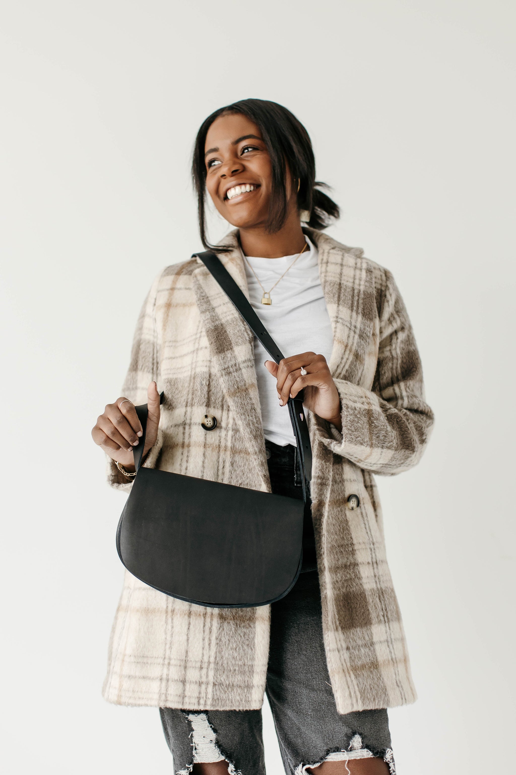 Smiling woman models a black leather crossbody purse and a stylish plaid overcoat.
