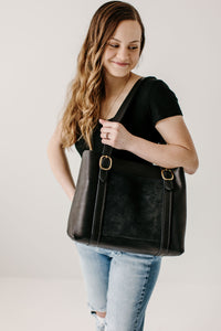 A woman models a stylish black leather tote bag with gold buckle details, wearing a black top and ripped jeans.
