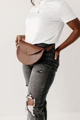 Woman models a stylish brown leather belt bag with distressed gray jeans and a white t-shirt against a white background.

