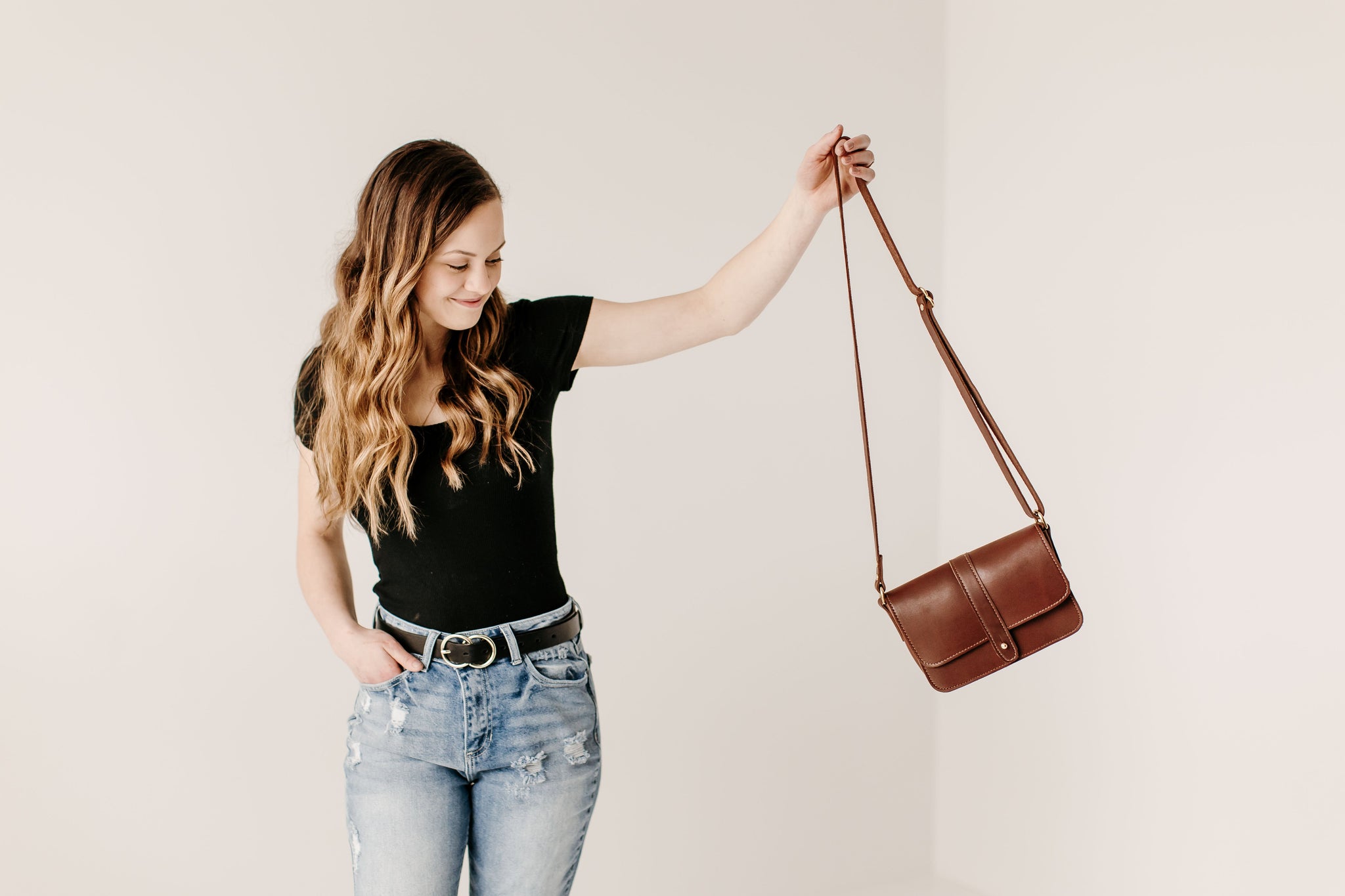 A stylish woman in jeans and a black top holds up a brown leather crossbody bag.
