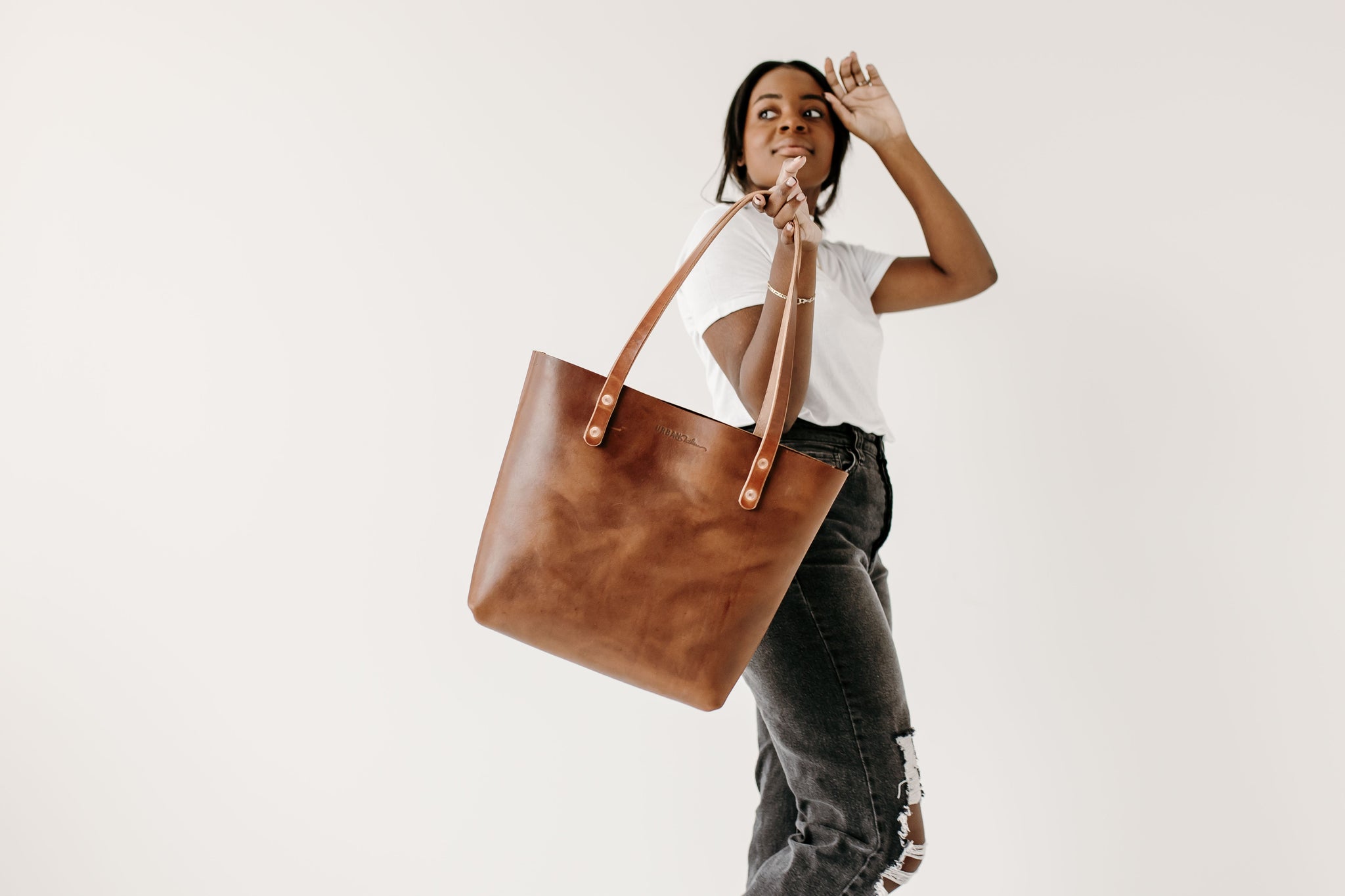 A Black woman models a brown leather tote bag, posing in front of a white background while holding the handbag.
