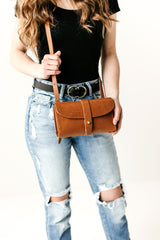 Woman holds a brown leather crossbody bag, showcasing its handmade design against a background of distressed denim jeans.
