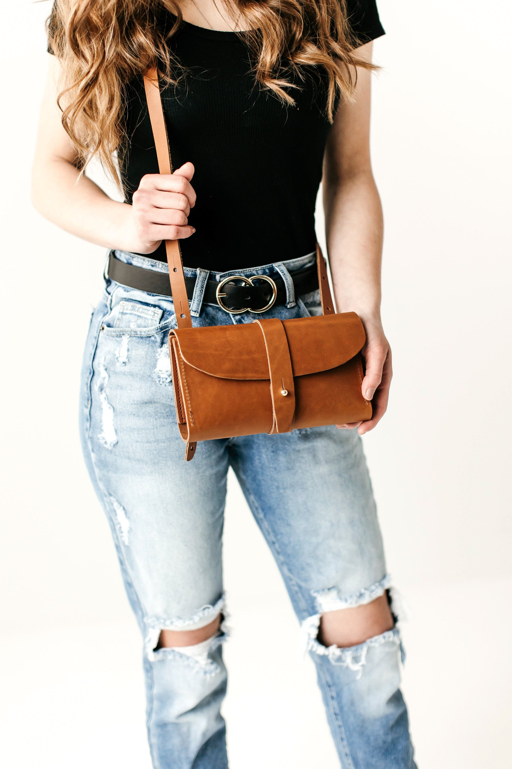 Woman holds a brown leather crossbody bag, showcasing its handmade design against a background of distressed denim jeans.
