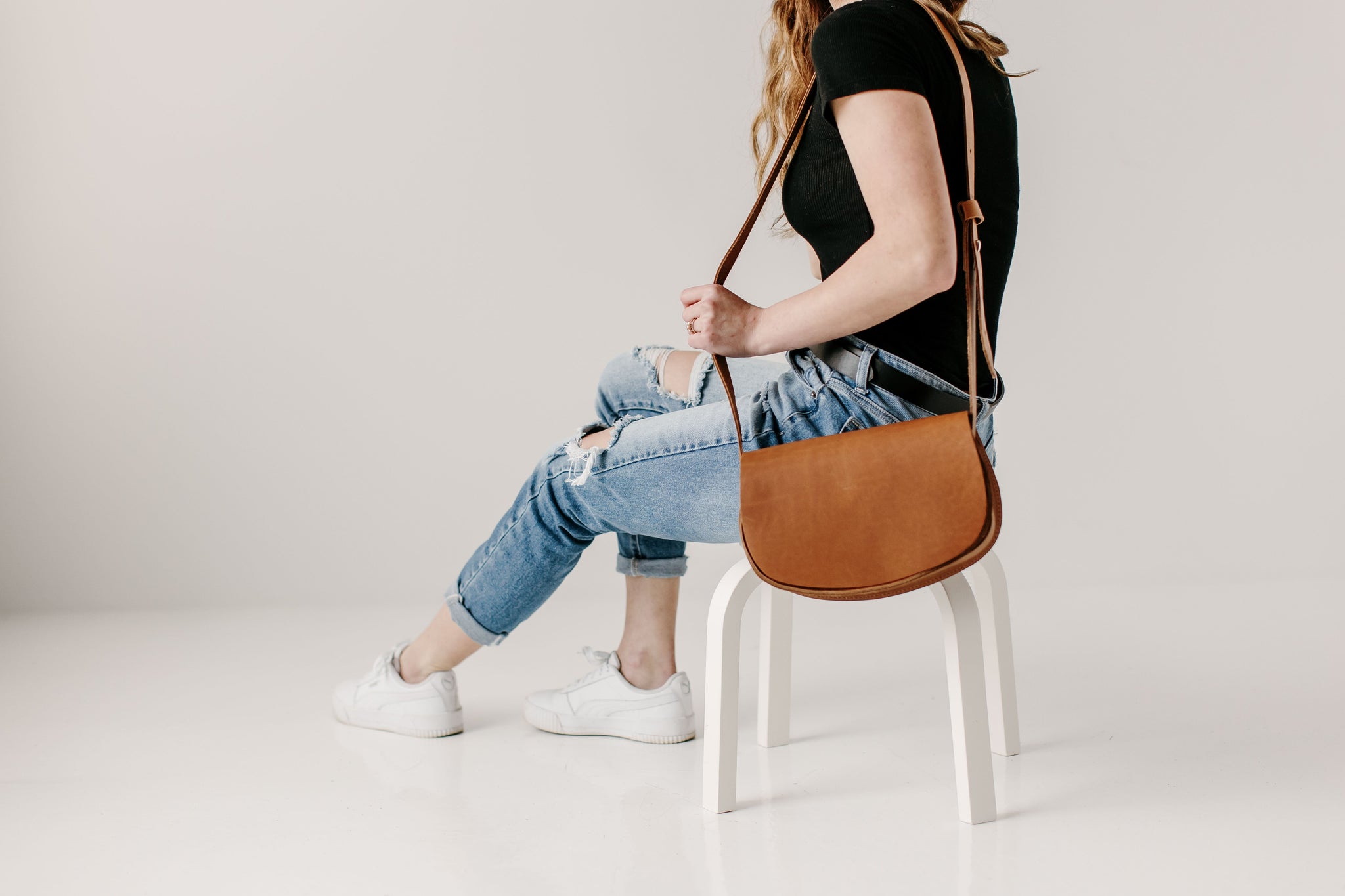 A woman models a brown leather crossbody bag while sitting on a white stool, wearing ripped jeans and white sneakers.
