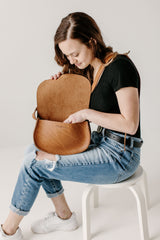 Woman checks inside her Urban Southern brown leather saddle bag while wearing ripped jeans and white sneakers.
