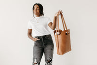 Woman models a stylish brown leather tote bag against a white background.
