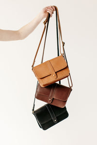 Hand displays a stack of three leather crossbody bags in tan, brown, and black against a white background.
