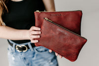 A woman holds two Urban Southern leather pouches, displaying their textured red leather and gold zippers.
