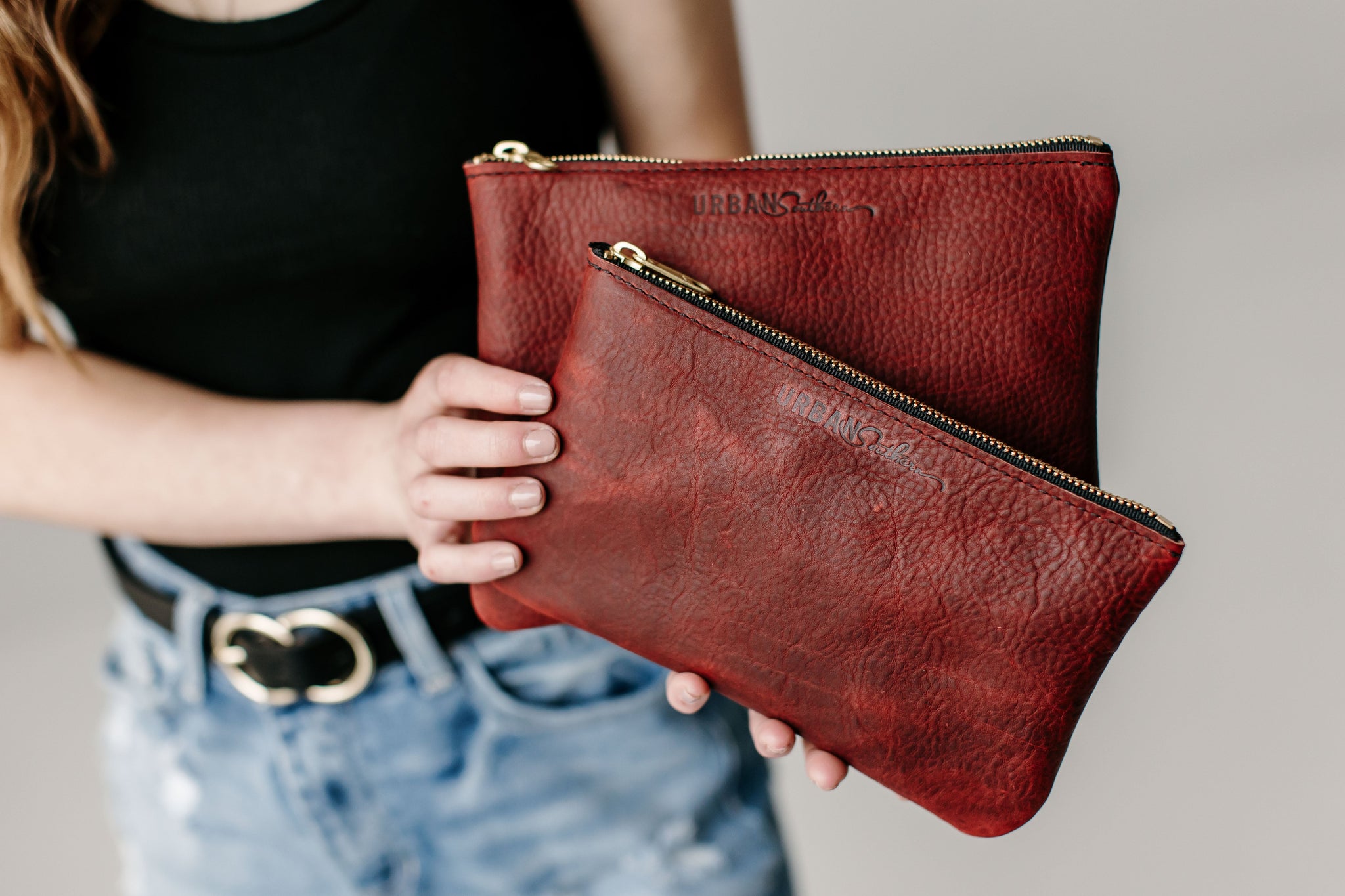 A woman holds two Urban Southern leather pouches, displaying their textured red leather and gold zippers.
