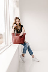 A woman in ripped jeans sits by a bright window, checking her stylish leather tote bag.
