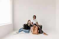 Two smiling women model stylish leather tote bags while sitting in a bright, minimalist studio.
