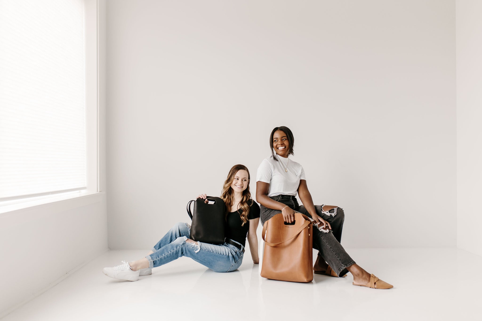 Two smiling women model stylish leather tote bags while sitting in a bright, minimalist studio.

