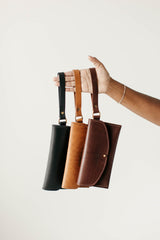 A hand models three stylish leather crossbody phone bags in black, tan, and brown against a white background.
