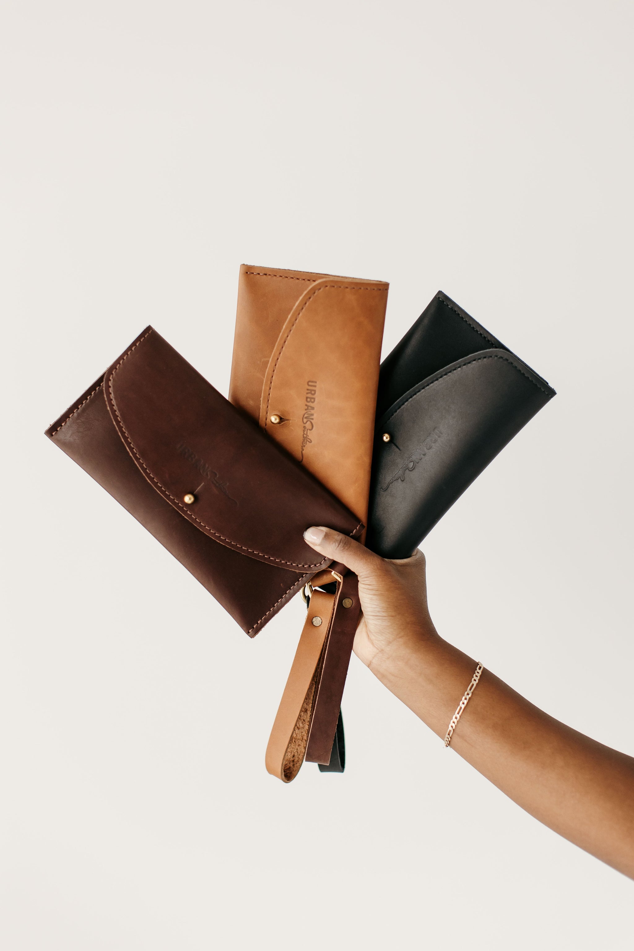 A hand displays three leather wallets in brown, tan, and black colors against a white background.
