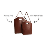 Two brown leather market totes, one full-sized and one mini, sit side-by-side against a white background.
