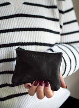 A woman wearing a striped sweater holds an Urban Collection black leather coin purse with burgundy nail polish.
