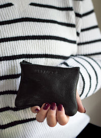A woman wearing a striped sweater holds an Urban Collection black leather coin purse with burgundy nail polish.
