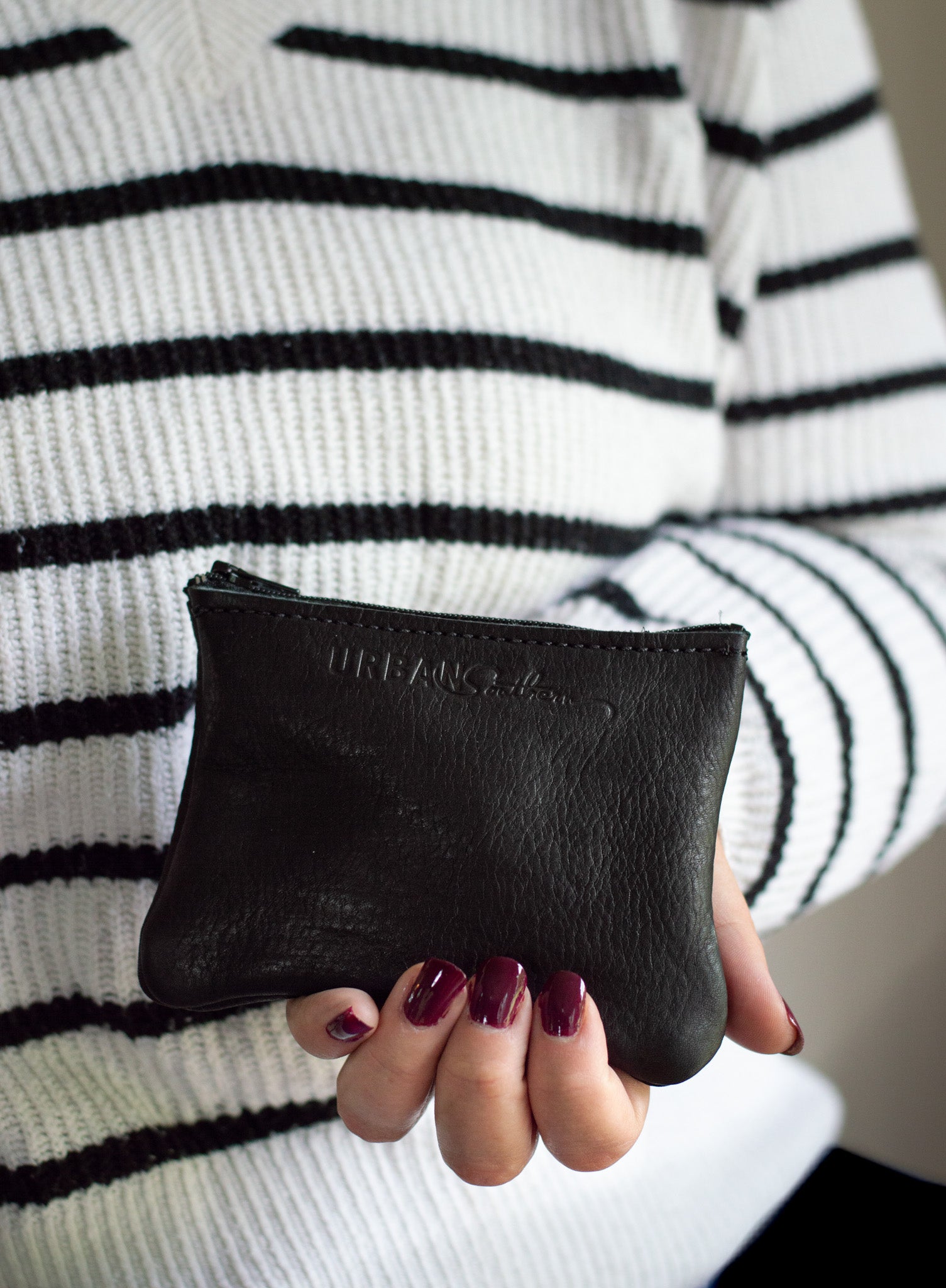 A woman wearing a striped sweater holds an Urban Collection black leather coin purse with burgundy nail polish.
