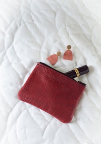 A red leather Urban Southern pouch sits beside rose-colored fern earrings and Revlon mascara on a white quilted surface.
