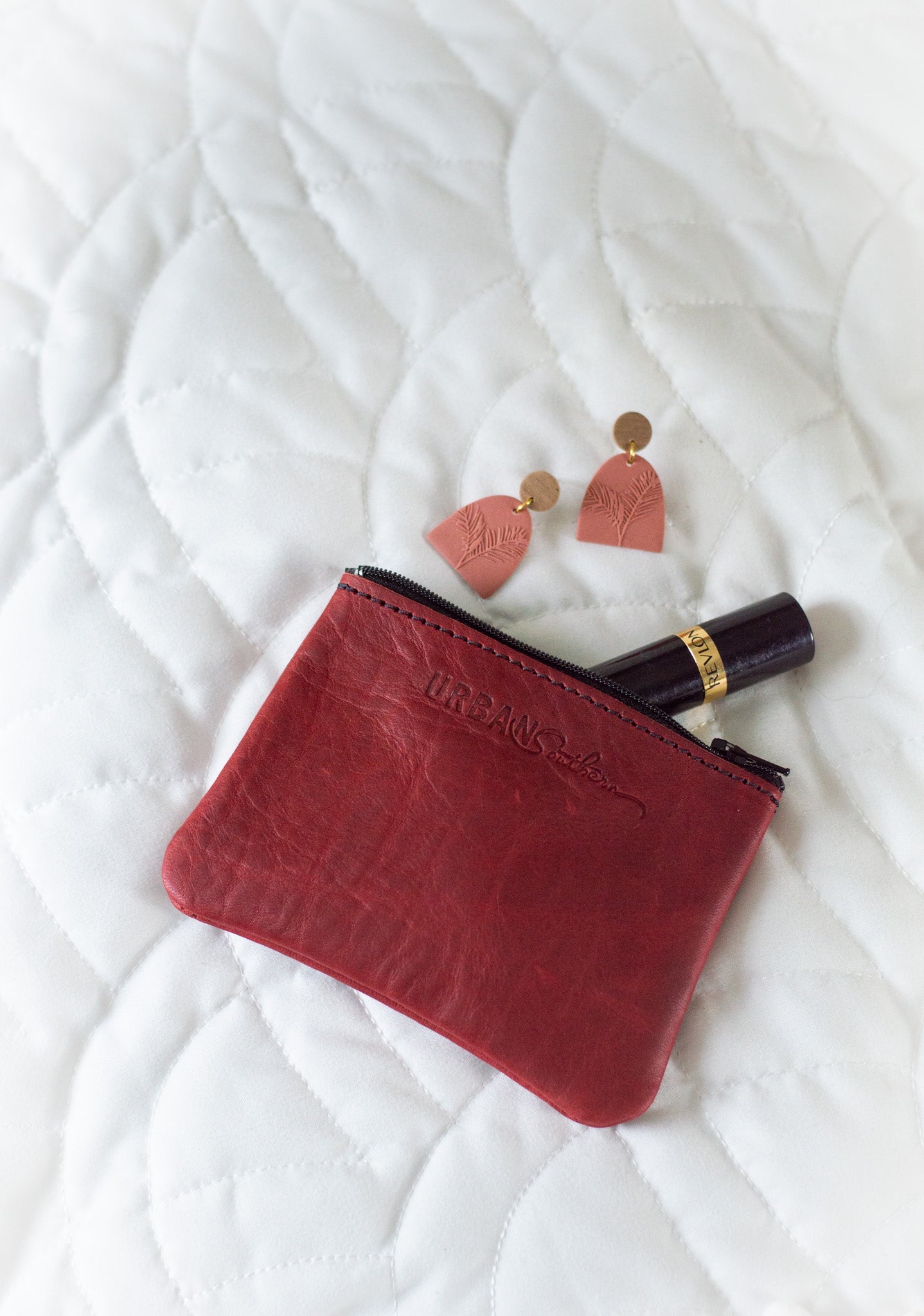 A red leather Urban Southern pouch sits beside rose-colored fern earrings and Revlon mascara on a white quilted surface.

