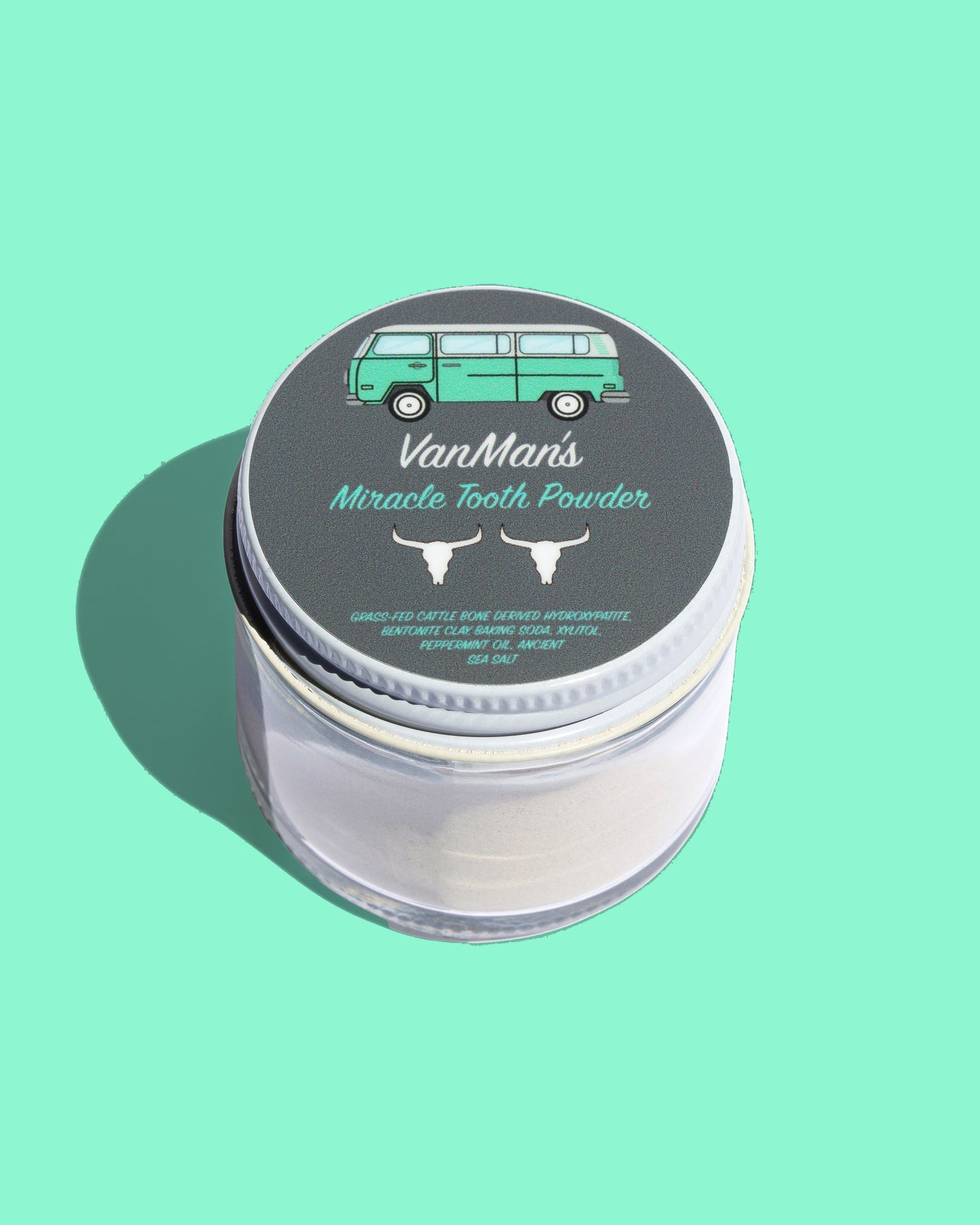 VanMan's Miracle Tooth Powder sits in a clear jar with a lid featuring a VW bus illustration and natural ingredients like bone-derived hydroxyapatite for healthy oral care.

