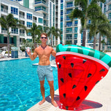 A fit man holds a watermelon pool float and a drink by a luxury pool on a sunny day.
