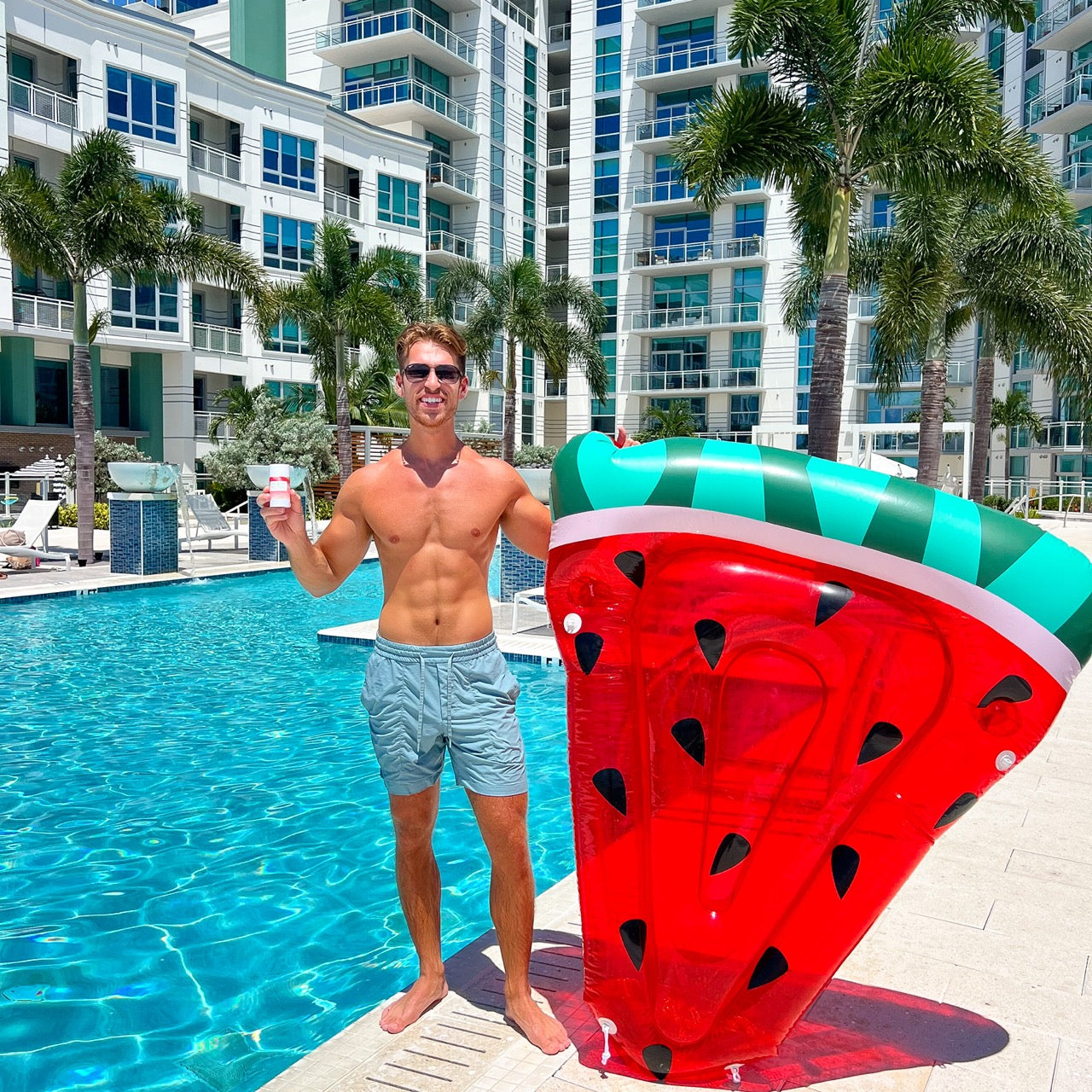 A fit man holds a watermelon pool float and a drink by a luxury pool on a sunny day.
