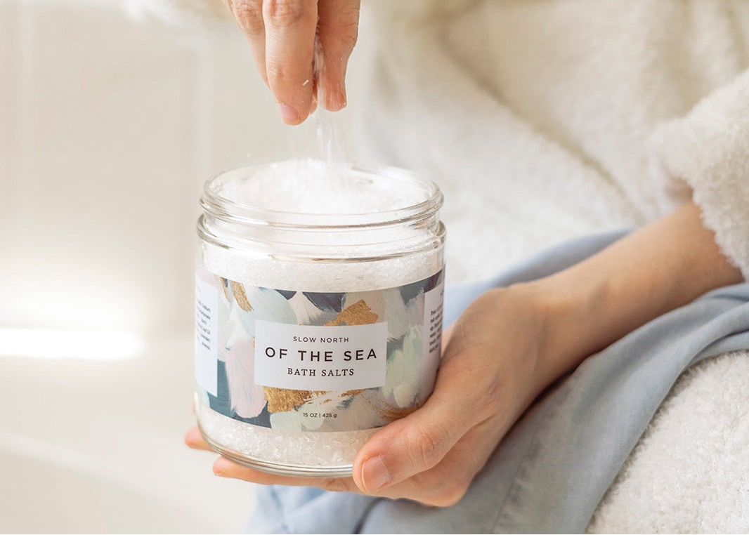 A person holding a glass jar of Slow North of the Sea Bath Salts, with their hand reaching in to scoop the white bath salts. The person is wearing a light blue sweatshirt and a white towel, set against a soft, warm, blurred background.