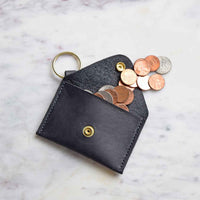 A handmade black leather coin purse with a keychain ring overflows with pennies and coins on a marble surface.
